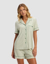 A woman with wavy blonde hair wears the Cozy Earth Women's Bamboo Stretch-Knit Short Sleeve Pajama Top in light green and white stripes, paired with matching shorts, standing against a plain light background and looking at the camera.|Color:Market Stripe in Julep
