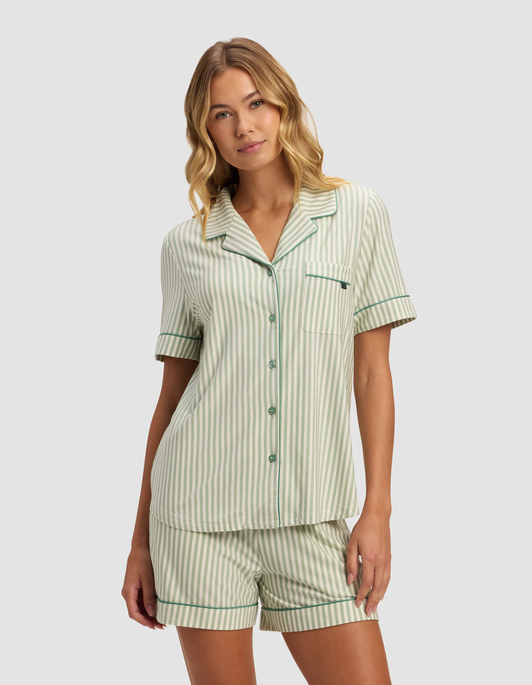 A woman with wavy blonde hair wears the Cozy Earth Women's Bamboo Stretch-Knit Short Sleeve Pajama Top in light green and white stripes, paired with matching shorts, standing against a plain light background and looking at the camera.|Color:Market Stripe in Julep