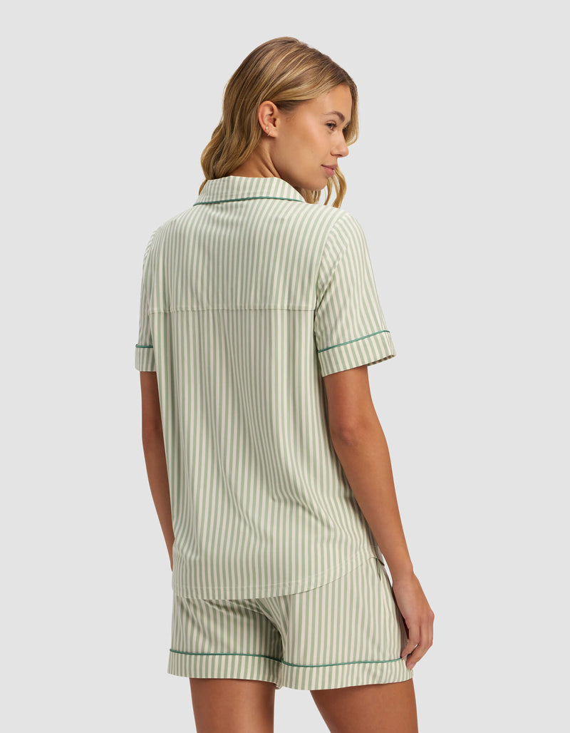 A woman with wavy blonde hair is seen from behind wearing the Cozy Earth Women's Bamboo Stretch-Knit Short Sleeve Pajama Top in light green and white stripes, paired with matching shorts featuring dark green trim, set against a plain background.