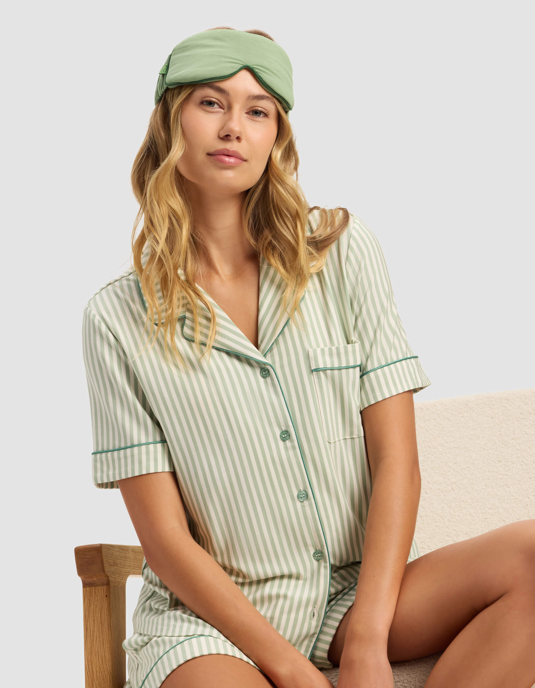 A woman with long blonde hair wears the Cozy Earth Women's Bamboo Stretch-Knit Short Sleeve Pajama Top, paired with a green sleep mask, as she sits calmly on a light-colored chair and looks at the camera.