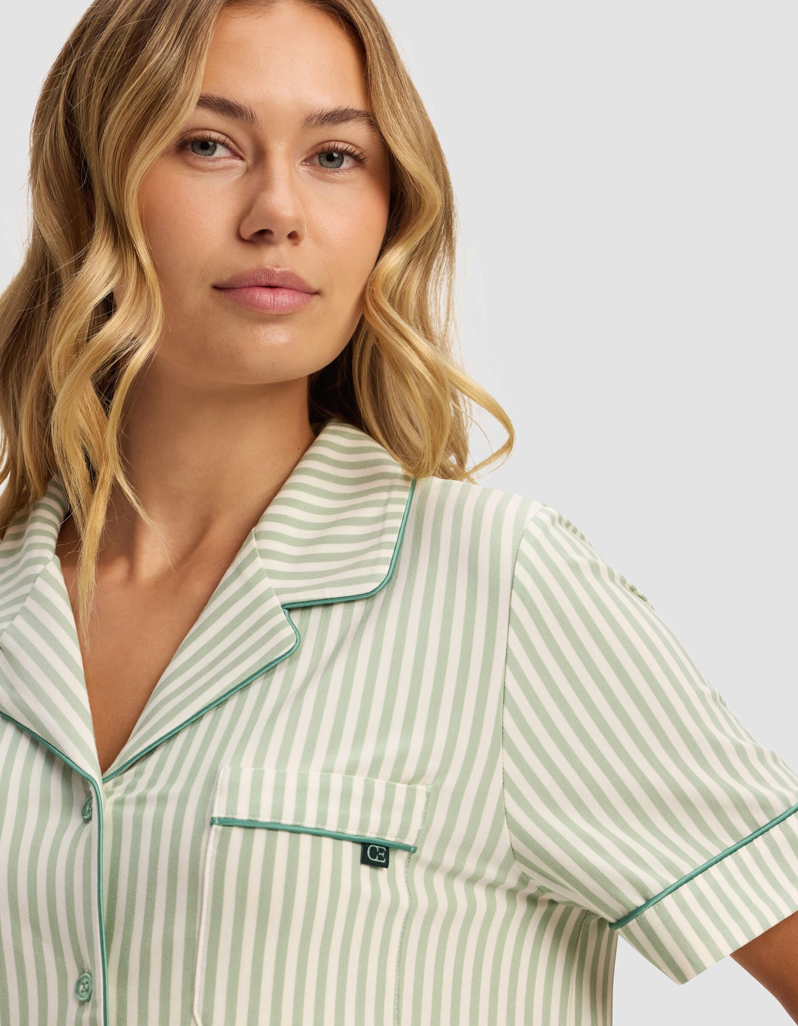 A woman with wavy blonde hair wears the Cozy Earth Women's Bamboo Stretch-Knit Short Sleeve Pajama Top, featuring a green and white striped design, collar, and front pocket, standing against a plain light background.
