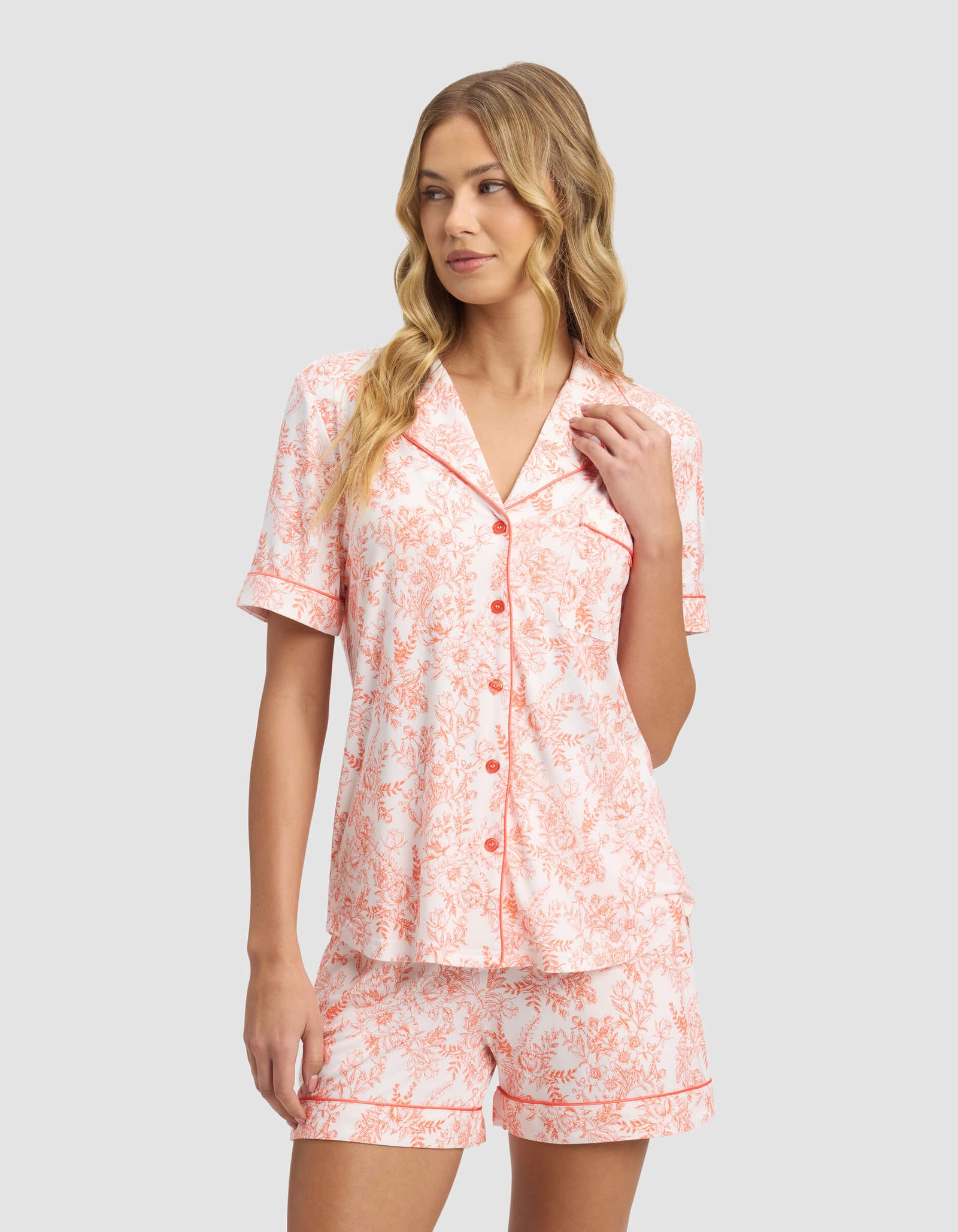 A woman with long, wavy blonde hair models the Cozy Earth Women's Bamboo Stretch-Knit Short Sleeve Pajama Top and matching shorts in a peach and white floral print, standing against a plain gray background.