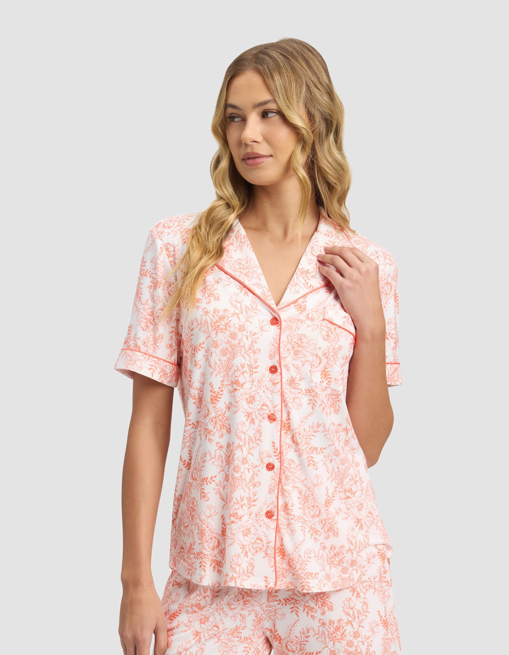 A woman wears the Cozy Earth Women's Bamboo Stretch-Knit Short Sleeve & Pant Pajama Set, featuring a pink and white floral pattern, as she stands against a light gray background and gazes slightly to the side.|Color:Melon Toile