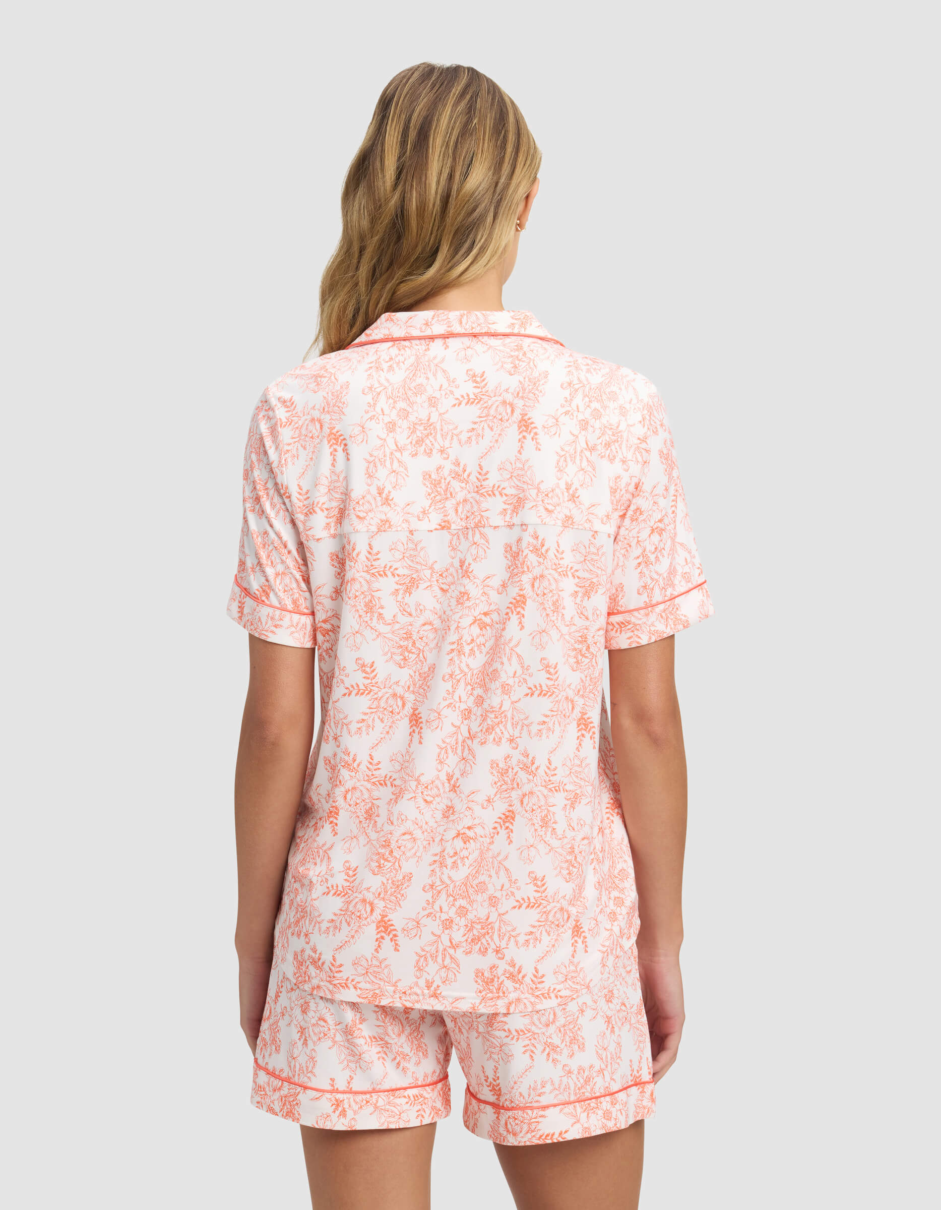 A woman with wavy blonde hair, seen from behind, wears the Cozy Earth Women's Bamboo Stretch-Knit Short Sleeve Pajama Top and matching shorts in a pink floral pattern on white.|Color:Melon Toile
