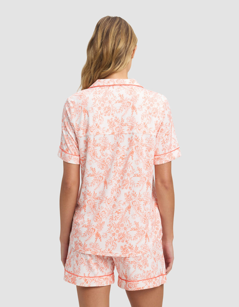 A woman with wavy blonde hair, seen from behind, wears the Cozy Earth Women's Bamboo Stretch-Knit Short Sleeve Pajama Top and matching shorts in a pink floral pattern on white.
