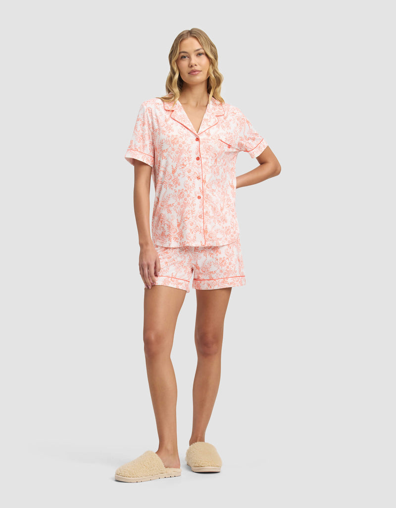 A woman wears the Cozy Earth Women's Bamboo Stretch-Knit Short Sleeve Pajama Top in light pink floral print with matching shorts and beige slippers, standing against a plain light gray background.
