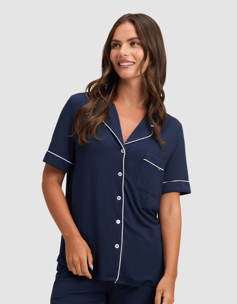 A woman smiles against a light gray background wearing HIDE's Women's Bamboo Stretch-Knit Short Sleeve Pajama Top in navy blue with white trim, paired with matching shorts.