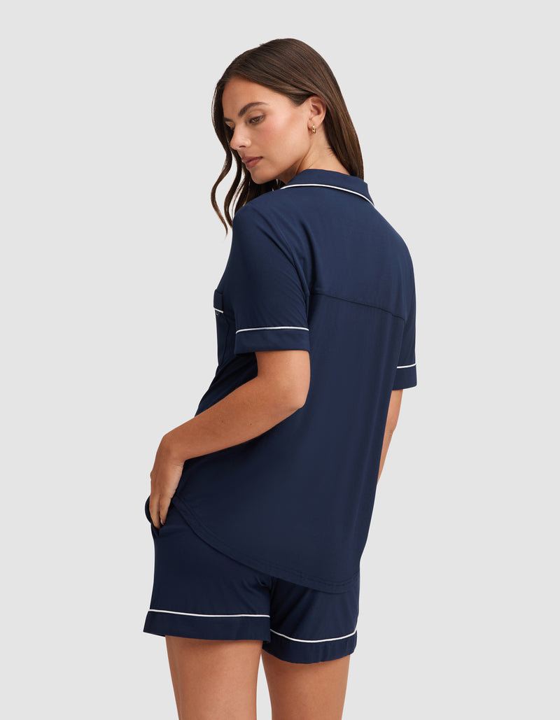 A woman wearing the HIDE Women's Bamboo Stretch-Knit Short Sleeve Pajama Top in navy blue with white piping faces away, glancing over her shoulder with a hand in her shorts pocket against a plain light background.
