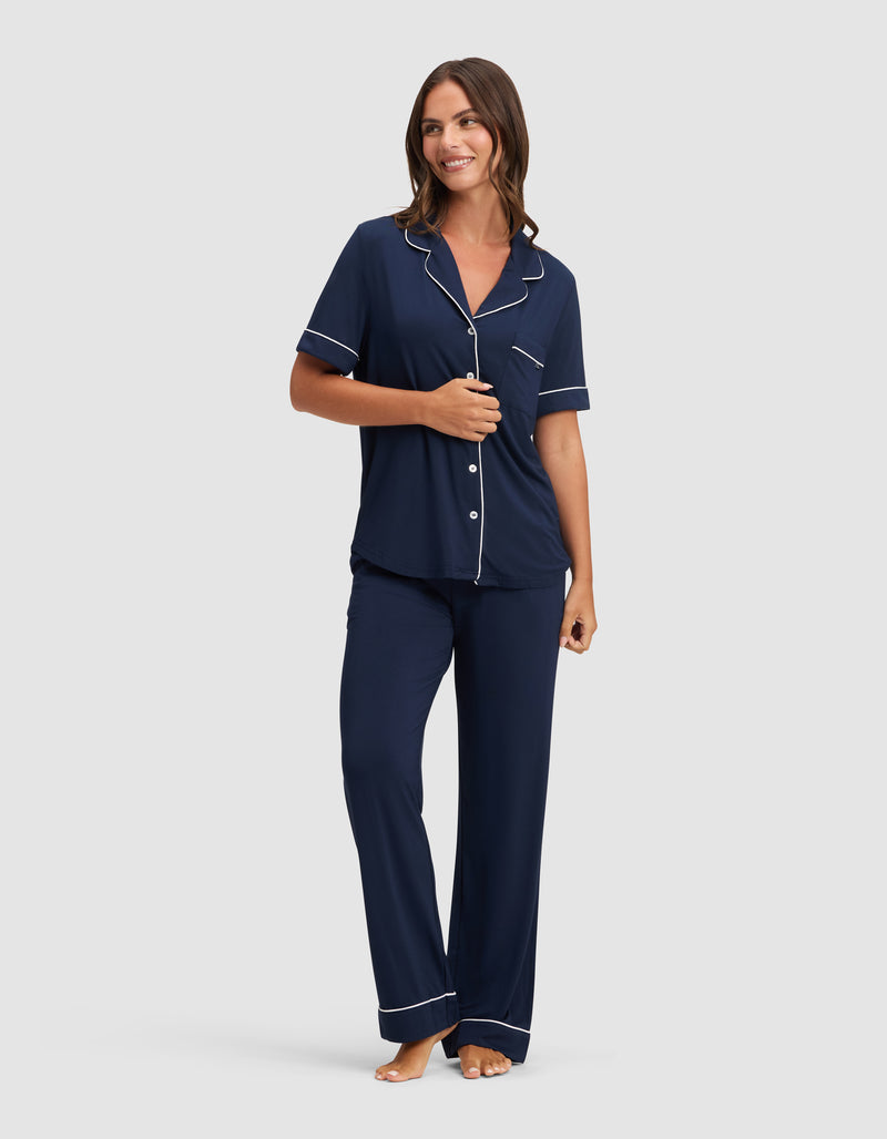 A woman smiles and looks to the side, barefoot and wearing HIDE's Women's Bamboo Stretch-Knit Short Sleeve Pajama Top in navy with white piping, paired with matching long pants.