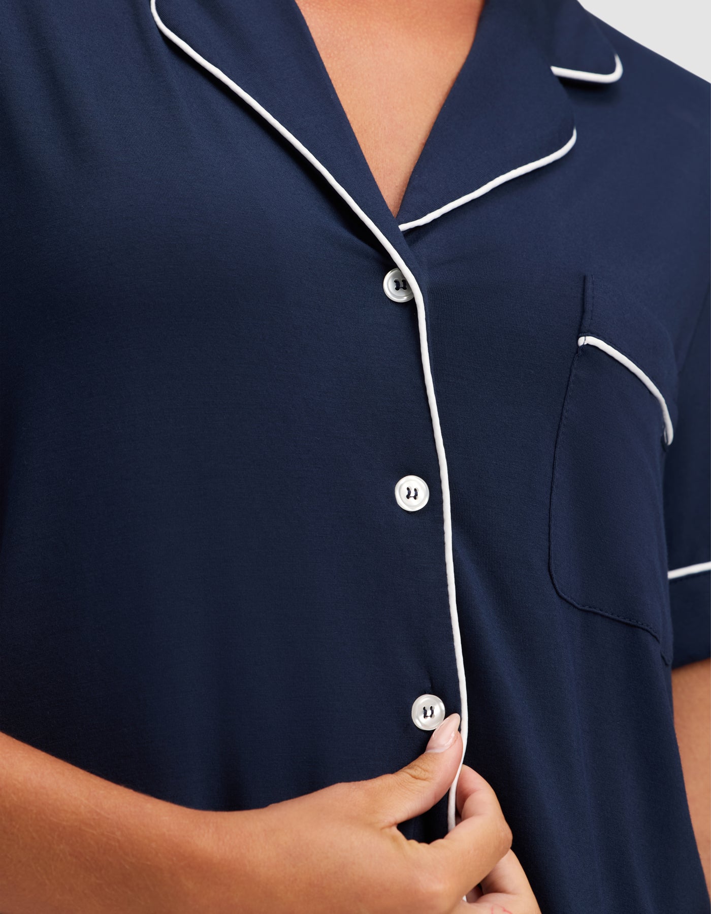 Close-up of a person wearing the HIDE Women's Bamboo Stretch-Knit Short Sleeve Pajama Top in navy blue with white piping, white buttons, and a chest pocket, holding a button with their right hand. 