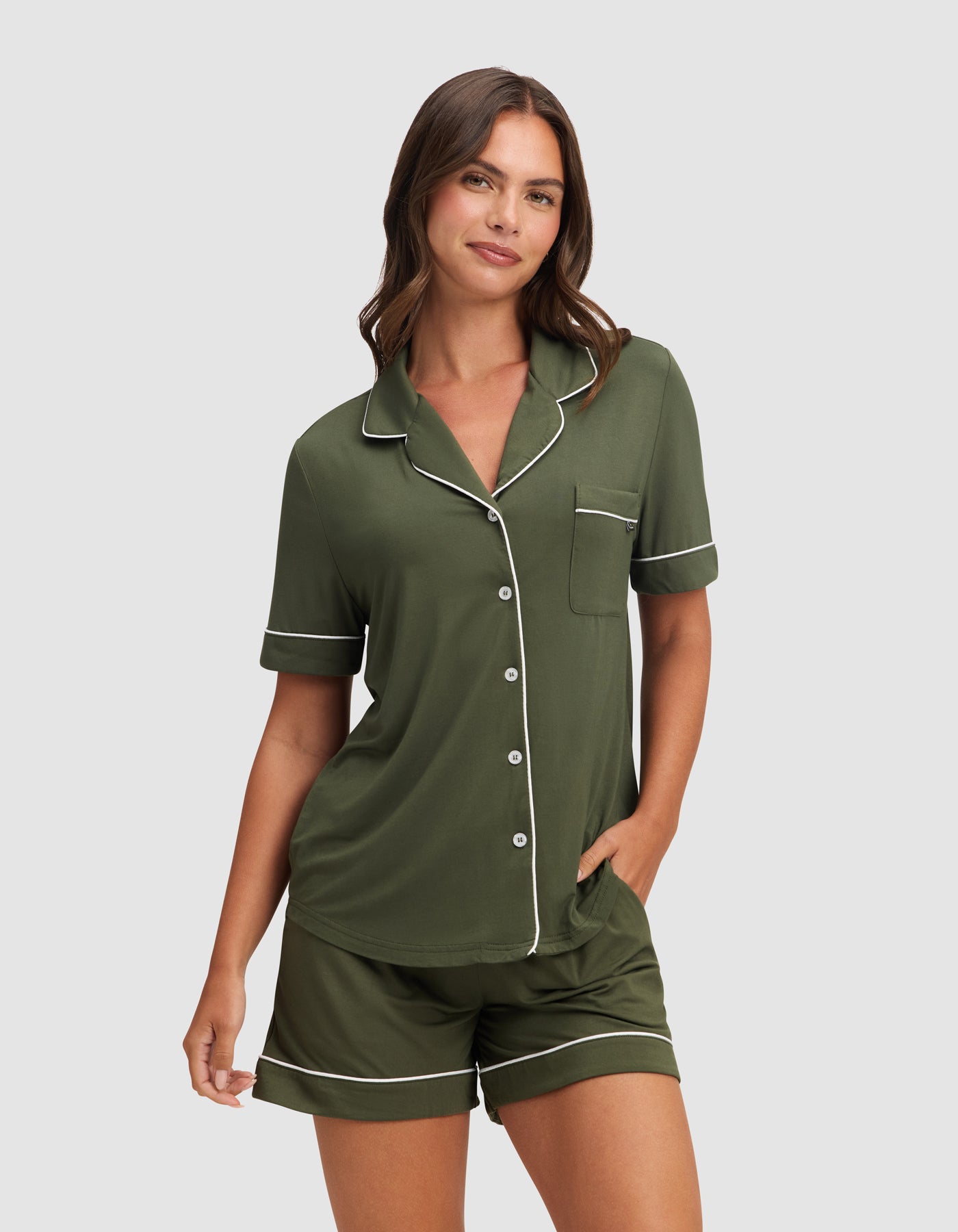 A woman stands against a plain background wearing the HIDE Women's Bamboo Stretch-Knit Short Sleeve Pajama Top in Olive, featuring white piping, front buttons, a chest pocket, and matching shorts with one hand in her pocket. 
