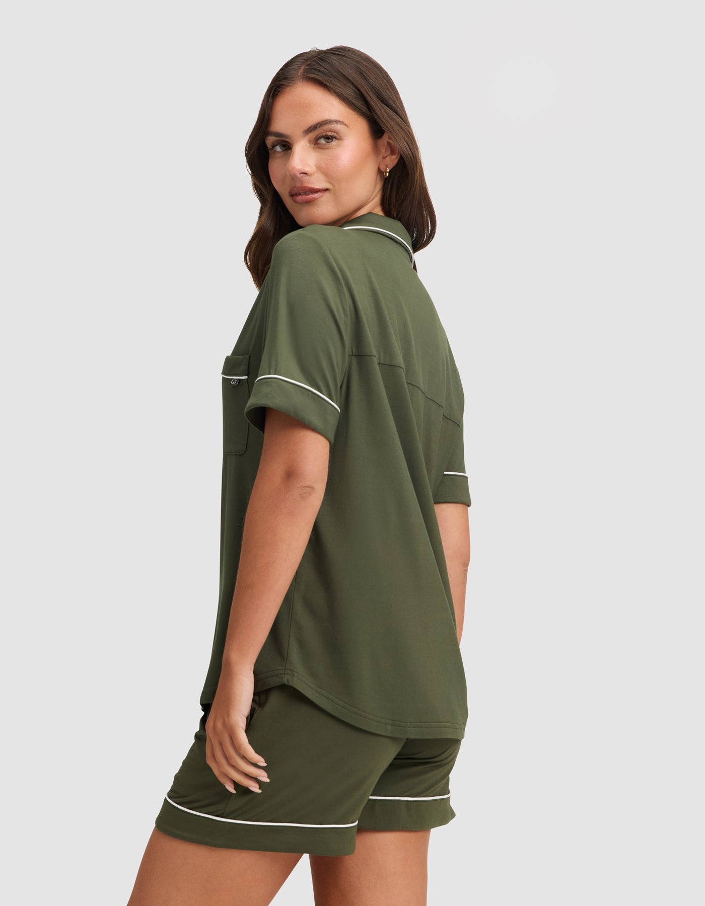 A woman with long brown hair wears the HIDE Women's Bamboo Stretch-Knit Short Sleeve Pajama Top in olive green with white piping, paired with matching shorts. She stands slightly turned, looking over her shoulder and smiling softly. 