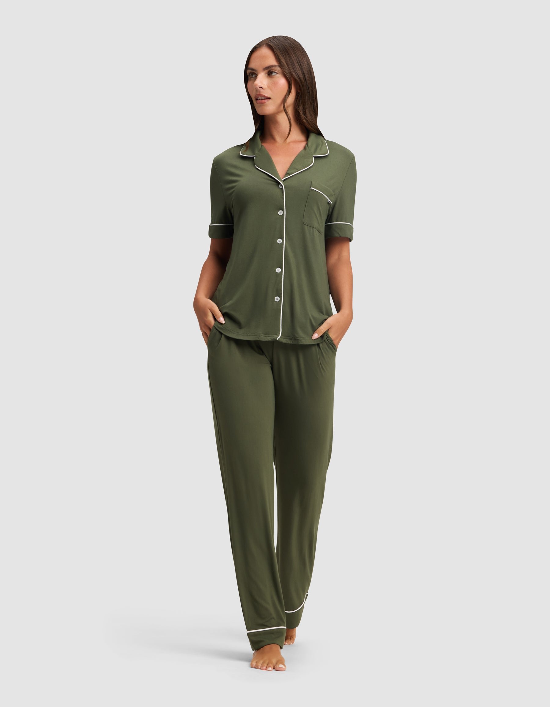 A woman stands barefoot against a plain background, hands in her pockets, wearing the HIDE Women's Bamboo Stretch-Knit Short Sleeve Pajama Top in olive green with white piping and button details. 