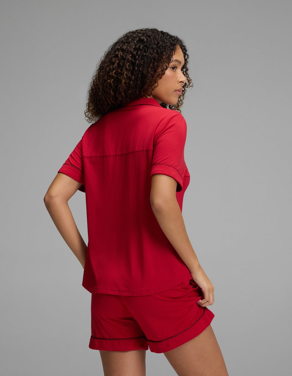 A person wears a red Women’s Bamboo Stretch-Knit Short Sleeve Pajama Set consisting of a short-sleeve button-down top with dark piping and matching shorts.