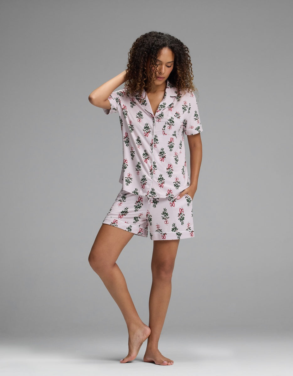 A woman wearing a Cozy Earth Women’s Bamboo Stretch-Knit Short Sleeve Pajama Set consisting of a short-sleeve button-down shirt and matching shorts in a pink holiday-themed print of bows and greenery.
