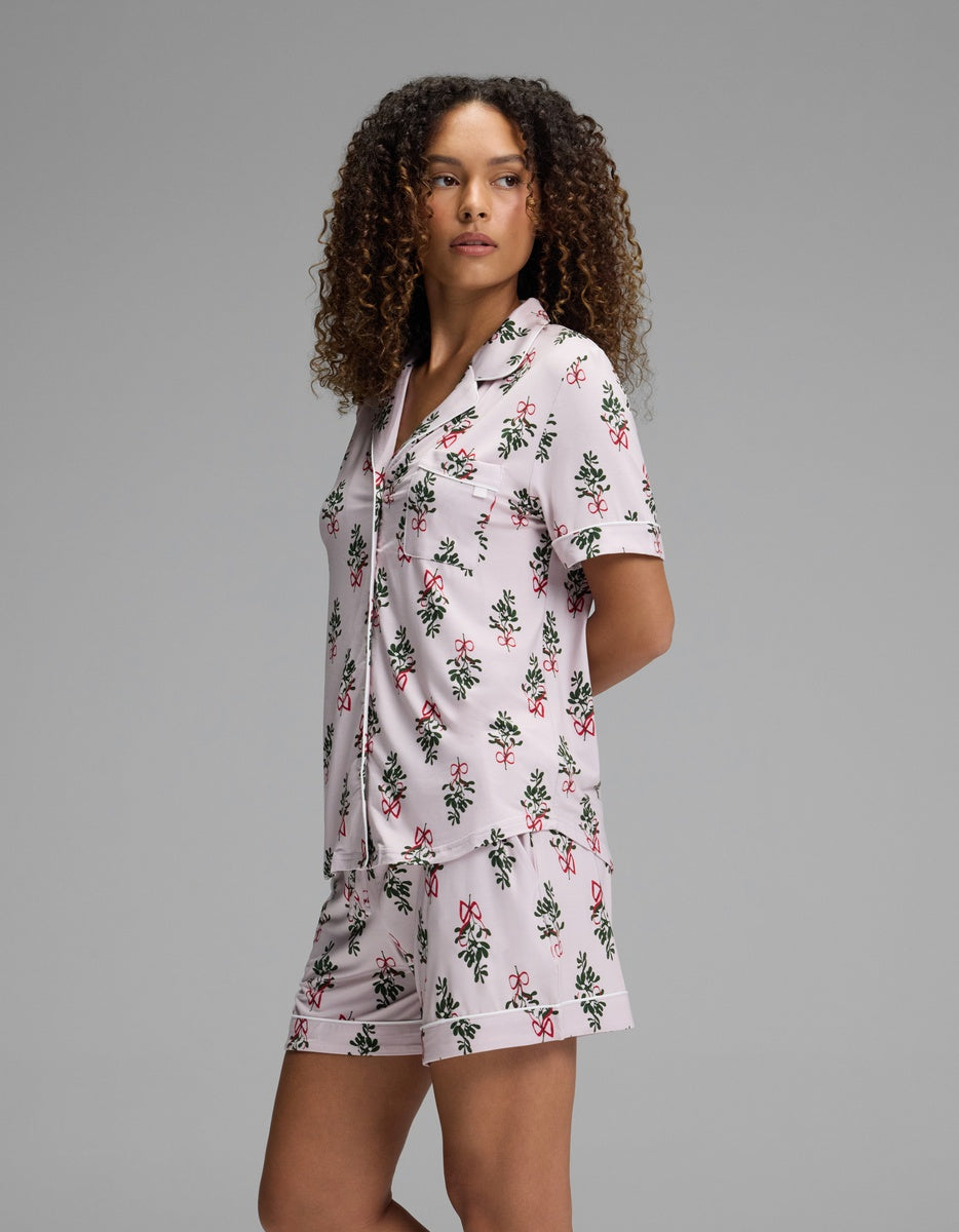 A woman wears a Cozy Earth Women’s Bamboo Stretch-Knit Short Sleeve Pajama Set, featuring a short-sleeve button-down shirt and matching shorts in a holiday-themed pattern against a pink background.