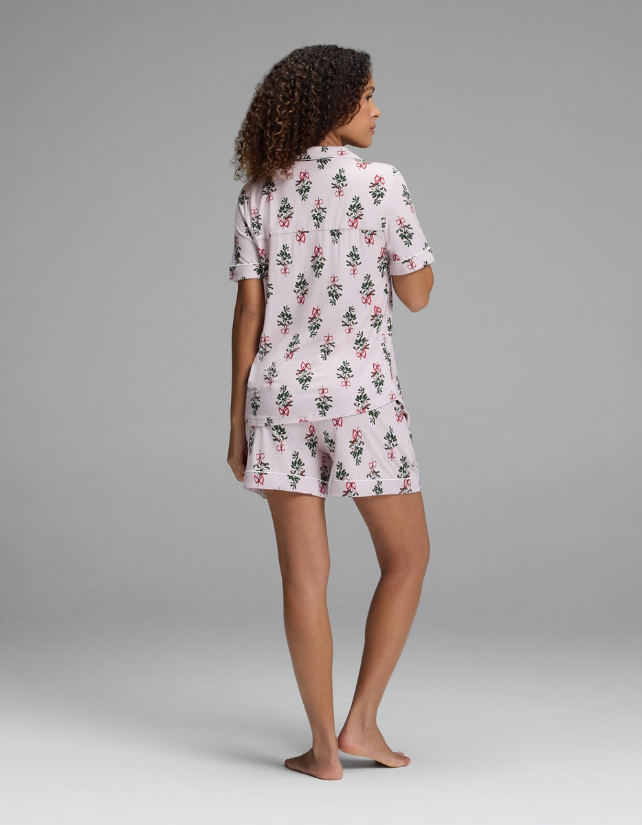 A woman seen from behind wearing a light pink Women’s Bamboo Stretch-Knit Short Sleeve Pajama Set featuring a pattern of green wreaths and red ribbons.