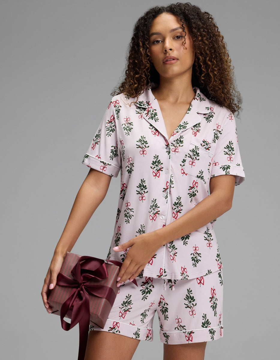 A woman wearing a Cozy Earth Women’s Bamboo Stretch-Knit Short Sleeve Pajama Set in a pink holiday-themed print of bows and greenery, holding a wrapped gift.