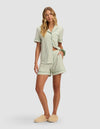 A woman models the Cozy Earth Women's Bamboo Stretch-Knit Short Sleeve Pajama Set in light green and white stripes with matching shorts, paired with beige slippers. She stands with one hand on her hip, her long wavy hair down.|Color:Market Stripe in Julep