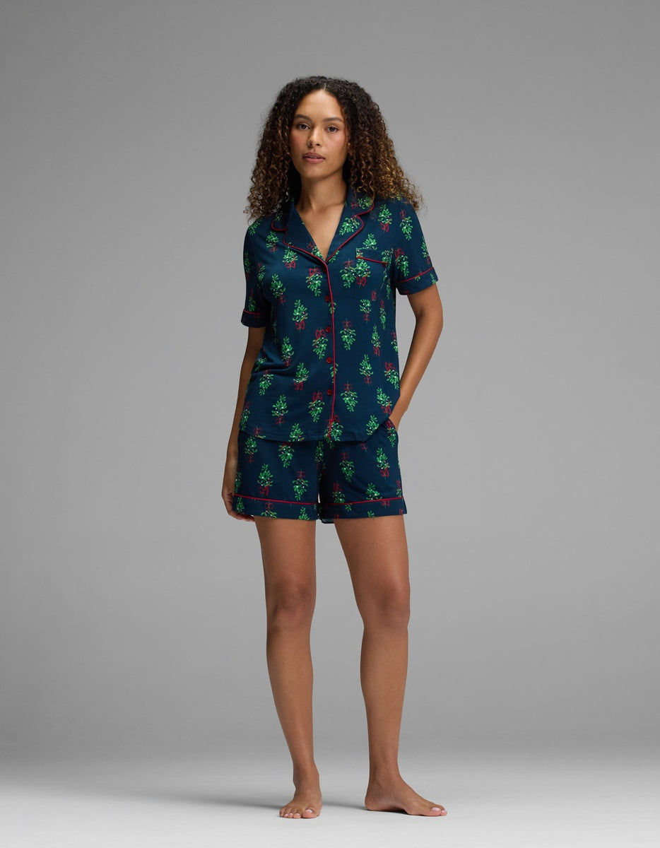 A woman wears a dark blue Cozy Earth Women’s Bamboo Stretch-Knit Short Sleeve Pajama Set, featuring a holiday-themed tree print, red piping, and a short-sleeved button-down top with matching shorts.