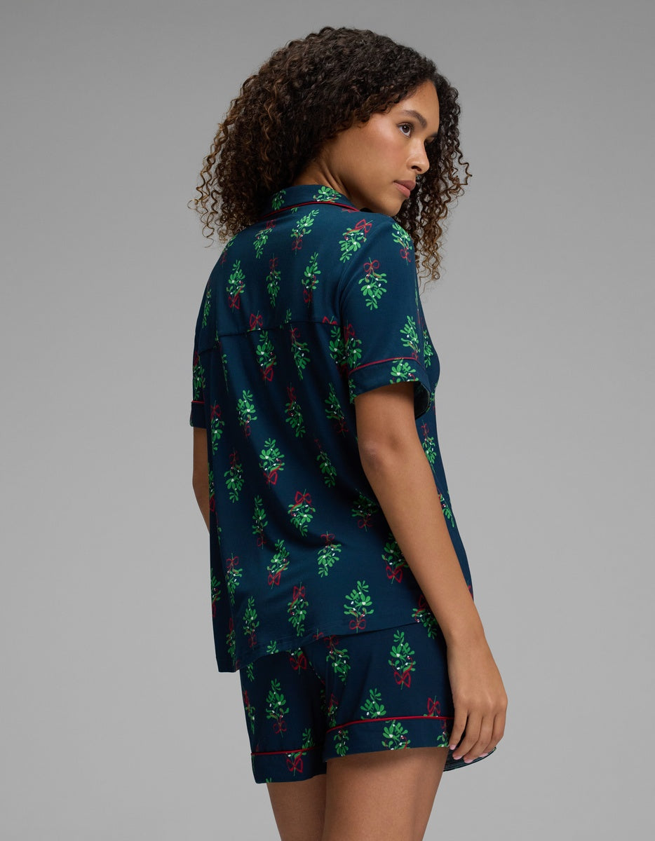 A woman wears a dark blue Cozy Earth Women’s Bamboo Stretch-Knit Short Sleeve Pajama Set featuring a green mistletoe and red bow print with red piping.