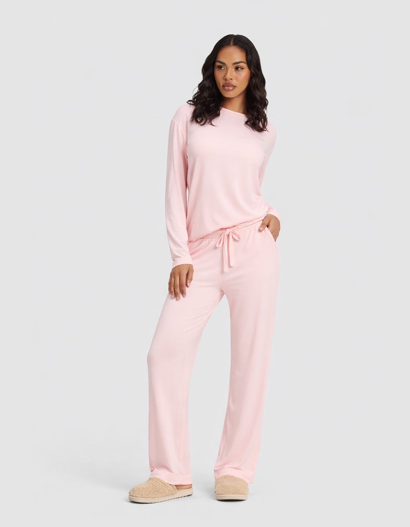 A woman wears the Cozy Earth Women's Bamboo Stretch-Knit Long Sleeve Lounge Tee & Pant Set in Blossom, paired with beige slippers, standing against a plain light gray background.|Color:Blossom