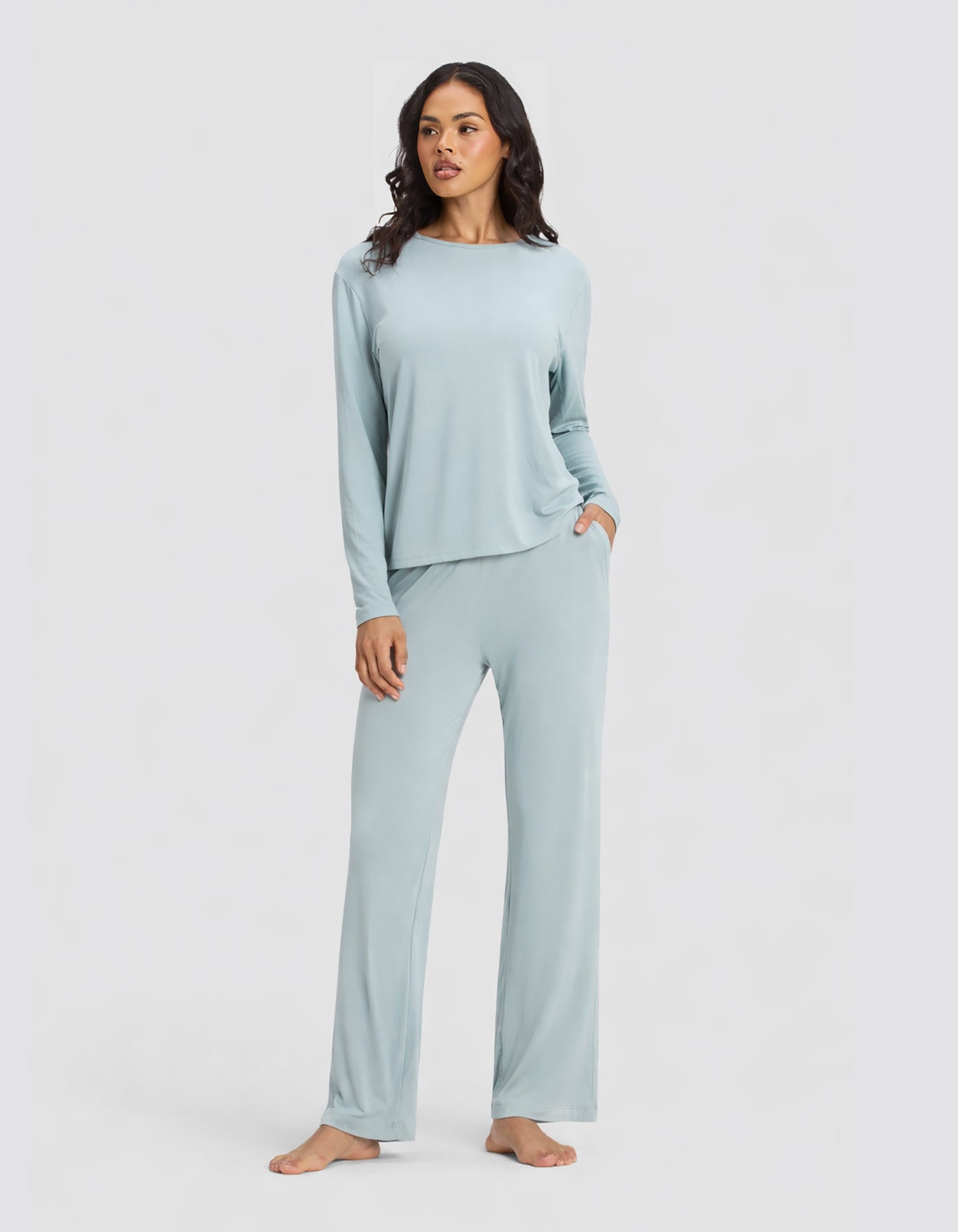 A woman stands barefoot against a plain background, wearing the Cozy Earth Women's Bamboo Stretch-Knit Long Sleeve Lounge Tee & Pant Set in Brook, with one hand in her pocket and a relaxed expression.