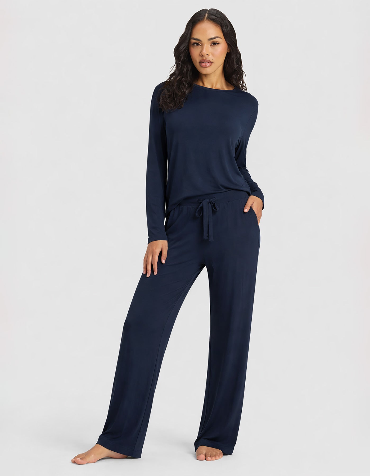 A woman poses barefoot in Cozy Earth's Women's Bamboo Stretch-Knit Long Sleeve Lounge Tee & Pant Set in navy, standing confidently against a plain light background.