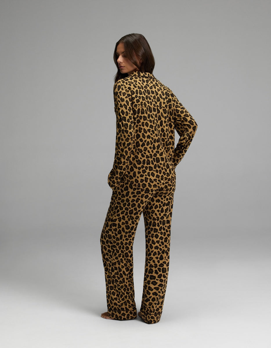 A person stands barefoot on a plain background, wearing the Cozy Earth Women’s Bamboo Stretch-Knit Long Sleeve Pajama Set in a matching leopard print, with their body turned slightly away and looking over one shoulder.