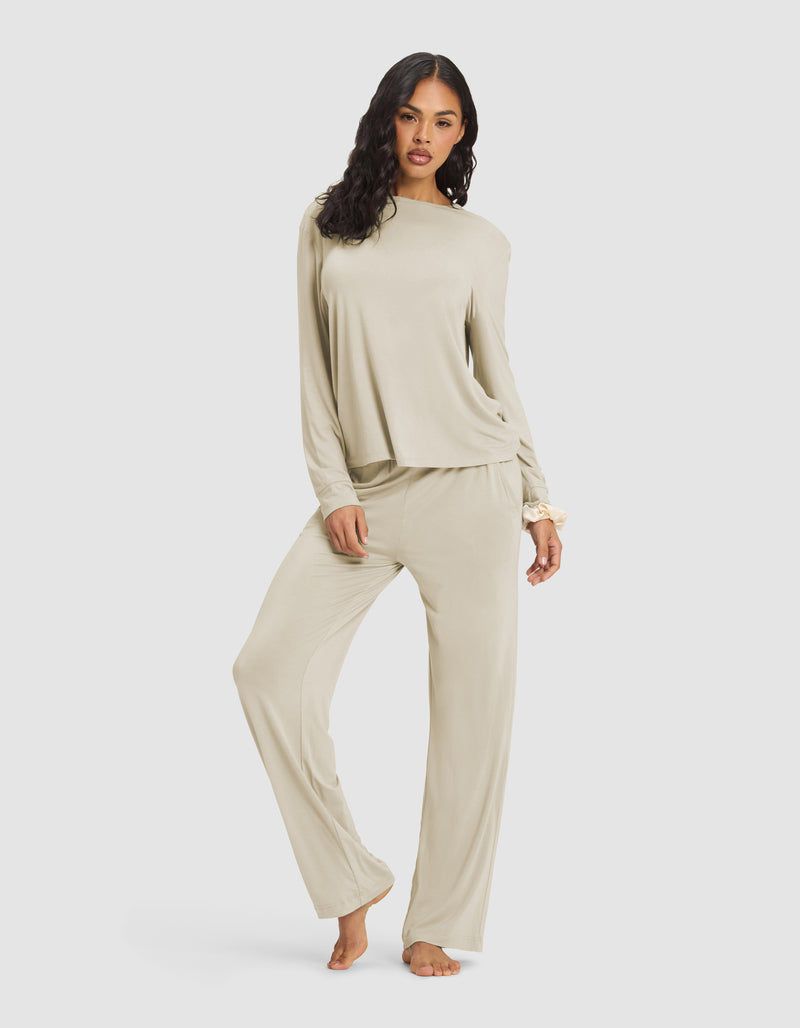 A woman with long, wavy dark hair models the Cozy Earth Women's Loews Hotel Lounge Set—a loose, light beige pajama set with long sleeves and wide-leg pants—as she stands barefoot against a plain white background.