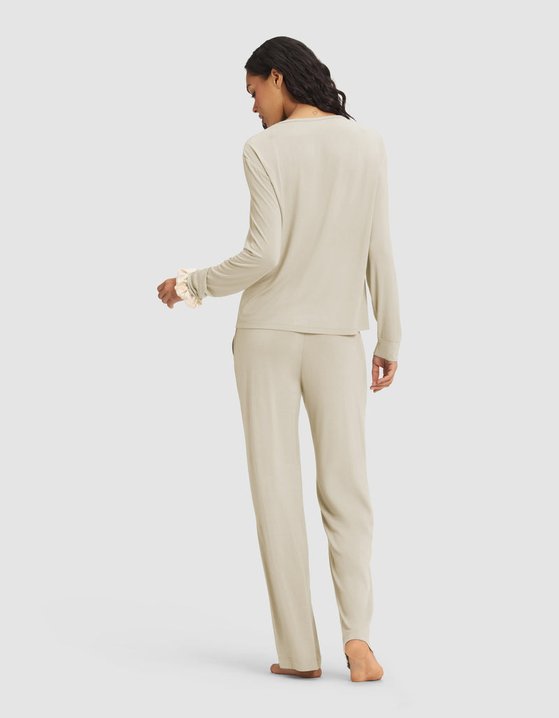 A woman with dark, wavy hair stands barefoot and facing away in the Cozy Earth Women's Loews Hotel Lounge Set—a light beige long-sleeve pajama top with matching pants—against a plain light gray background.