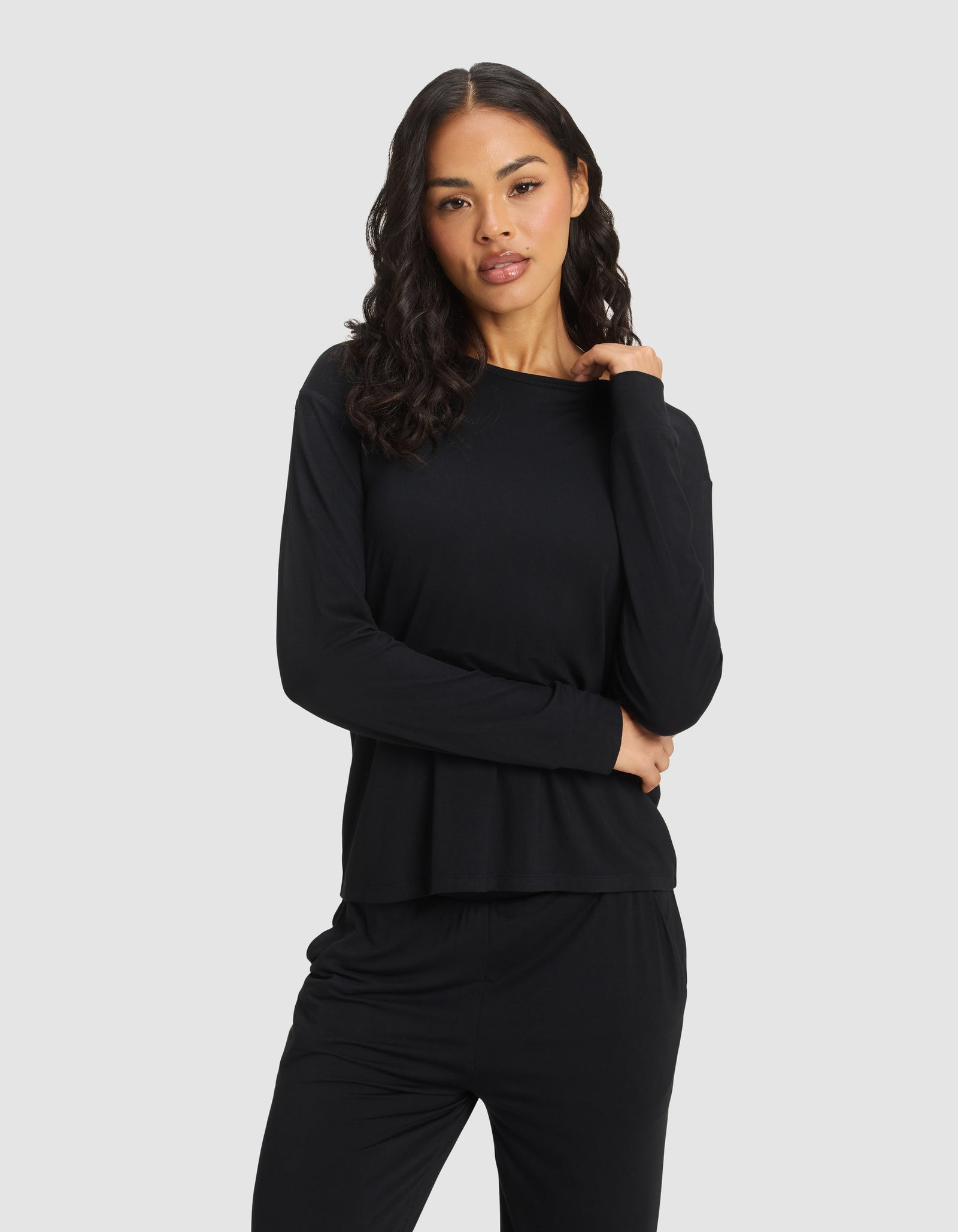 A woman with wavy dark hair models the Cozy Earth Women's Bamboo Stretch-Knit Long Sleeve Lounge Tee in black, standing against a plain light gray background with one hand on her shoulder and the other crossing her waist. 