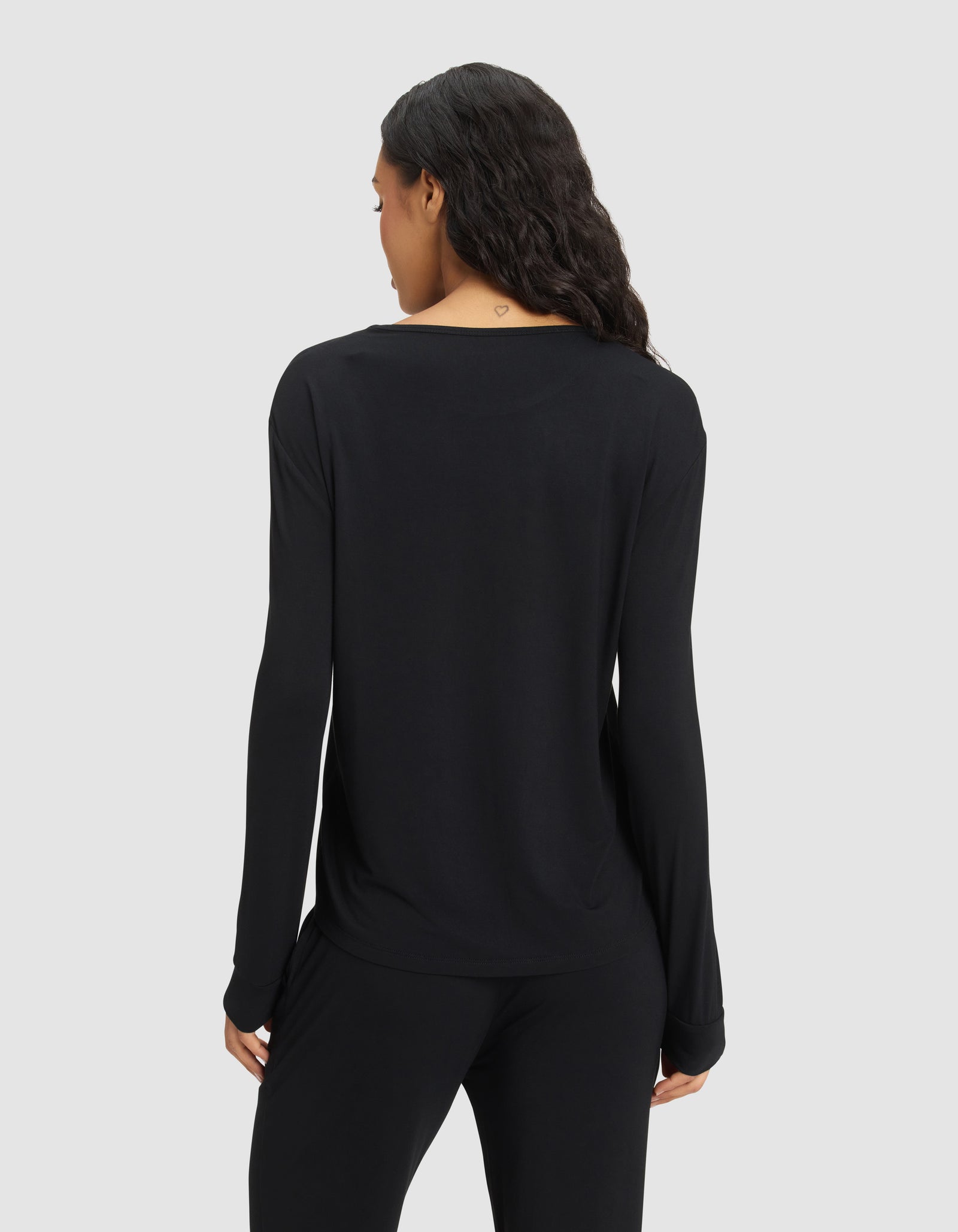 A woman with long dark hair is seen from behind wearing the Cozy Earth Women's Bamboo Stretch-Knit Long Sleeve Lounge Tee in black, paired with matching pants, standing against a plain light gray background. 