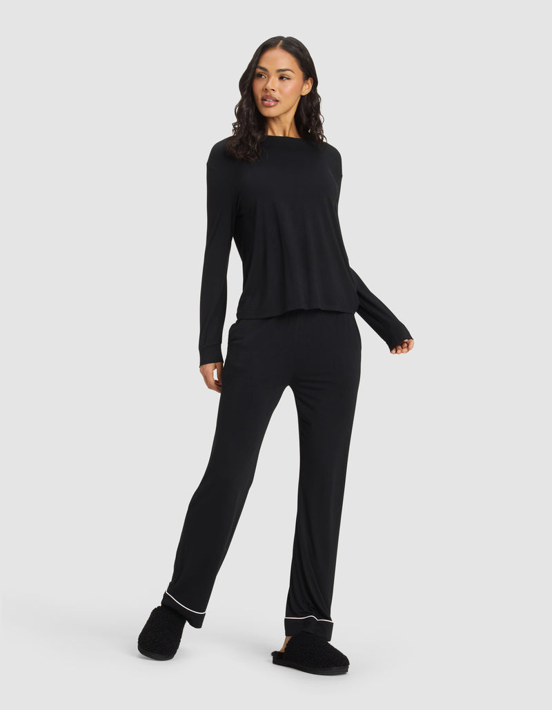 A woman stands against a plain background wearing the Cozy Earth Women's Bamboo Stretch-Knit Long Sleeve Lounge Tee in black and matching slippers, smiling slightly with one hand on her hip and looking to the side.