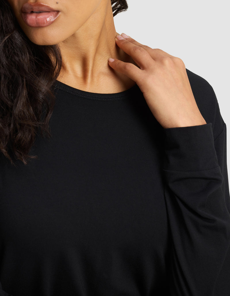 A woman with wavy dark hair wears the Women's Bamboo Stretch-Knit Long Sleeve Lounge Tee by Cozy Earth. She gently touches her neck with her right hand, and only the lower half of her face and upper torso are visible.