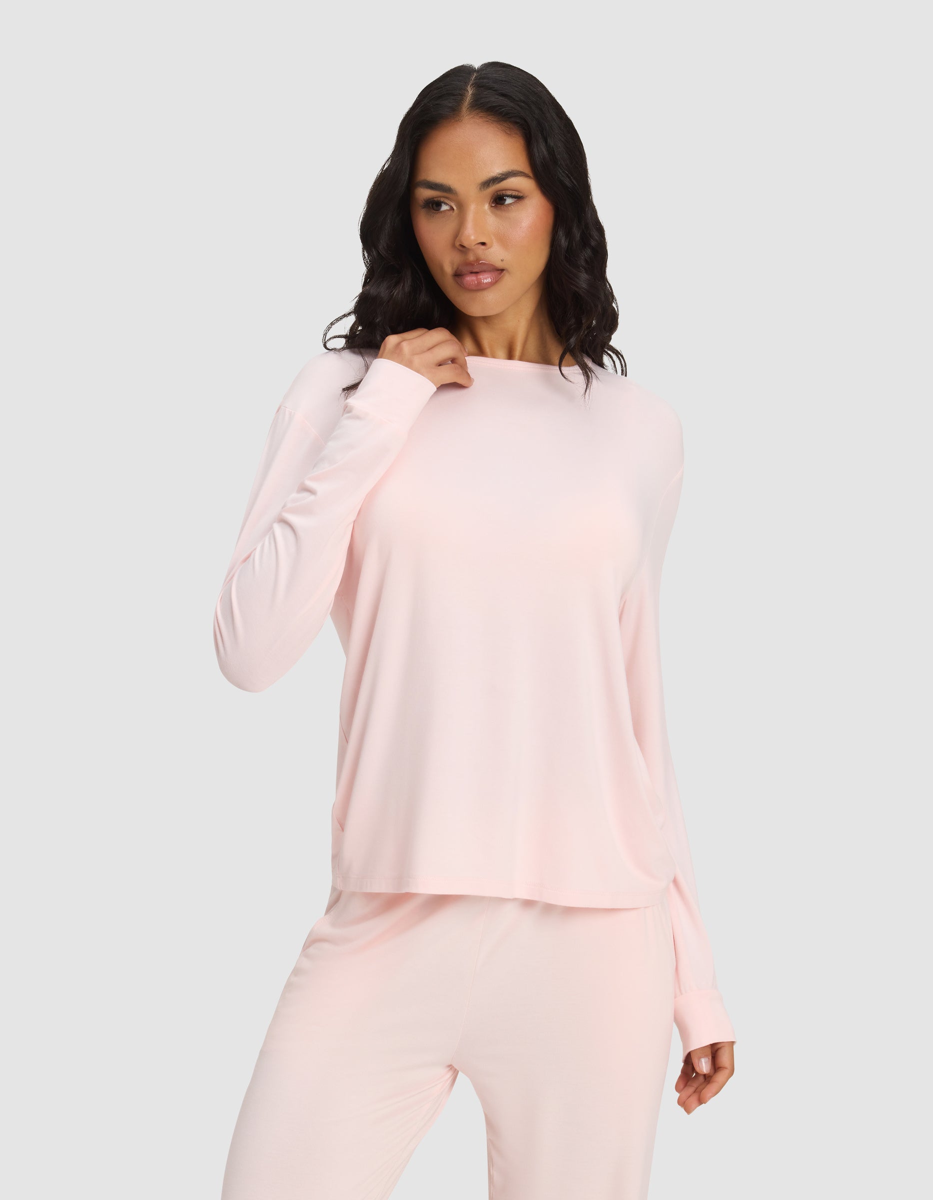 A woman with long, wavy dark hair wears the Cozy Earth Women's Bamboo Stretch-Knit Long Sleeve Lounge Tee in light pink, paired with matching pants, posing against a plain light gray background.|Color:Blossom