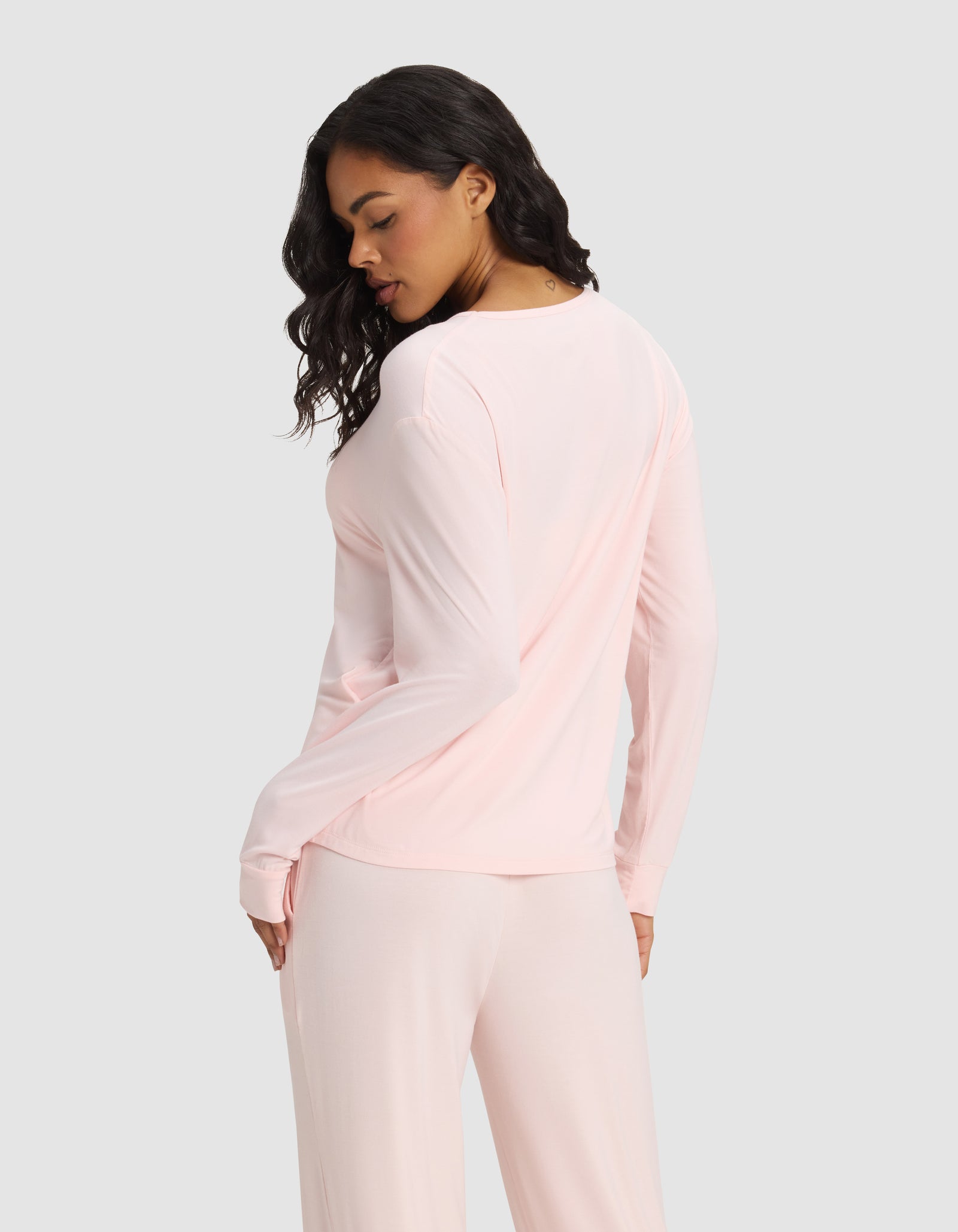 A woman with long dark hair wears the Cozy Earth Women's Bamboo Stretch-Knit Long Sleeve Lounge Tee in light pink, paired with matching pants, standing with her back turned and head slightly tilted against a plain light gray background.