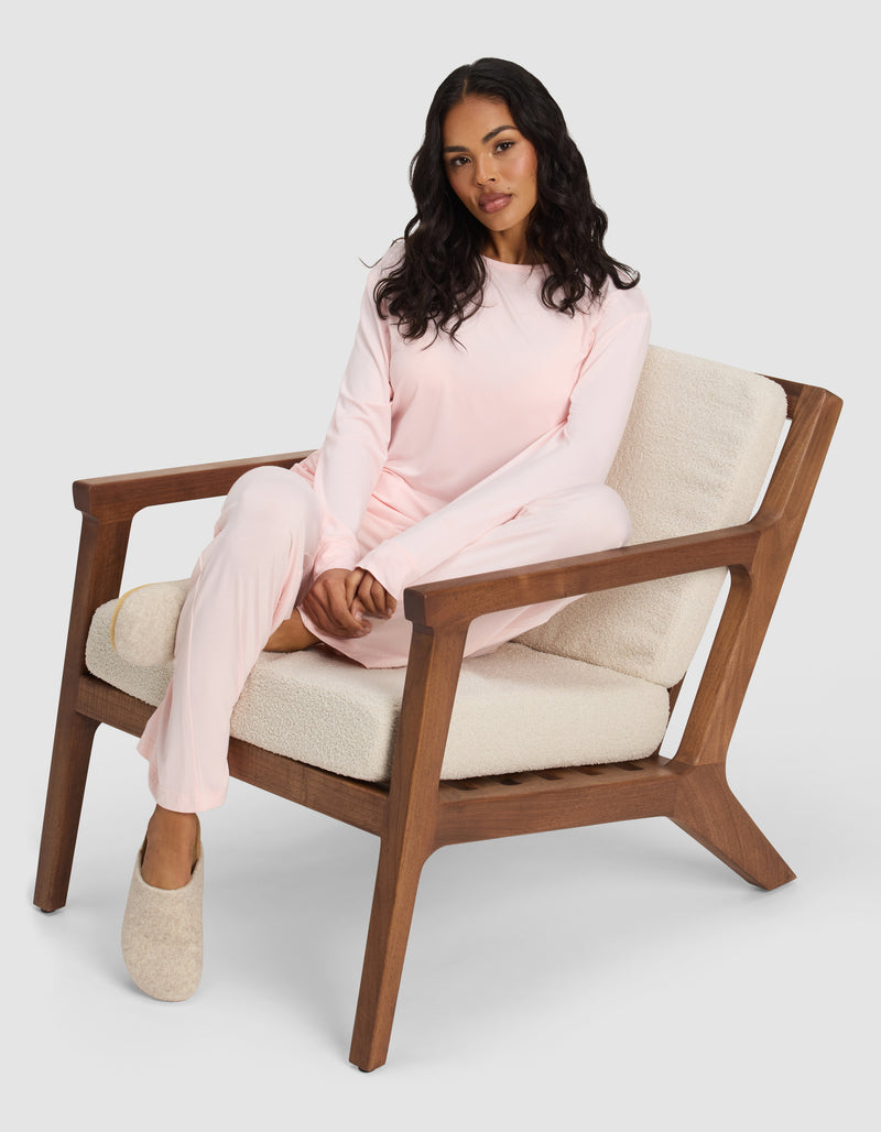 A woman with long dark hair sits on a cushioned wooden chair, wearing Cozy Earth's Women's Bamboo Stretch-Knit Long Sleeve Lounge Tee in light pink and beige slippers, her left leg crossed over her right knee against a plain background.