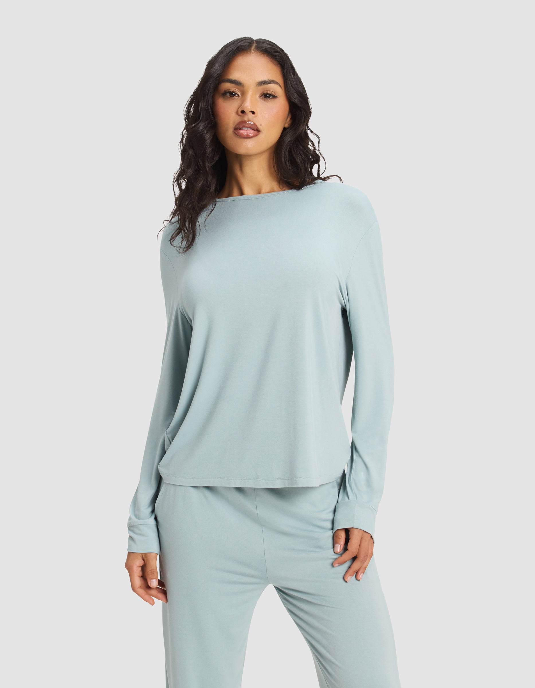 A woman with long, wavy hair wears the Cozy Earth Women's Bamboo Stretch-Knit Long Sleeve Lounge Tee in light blue, paired with matching pants. She stands against a plain light gray background, facing forward with a neutral expression.|Color:Brook