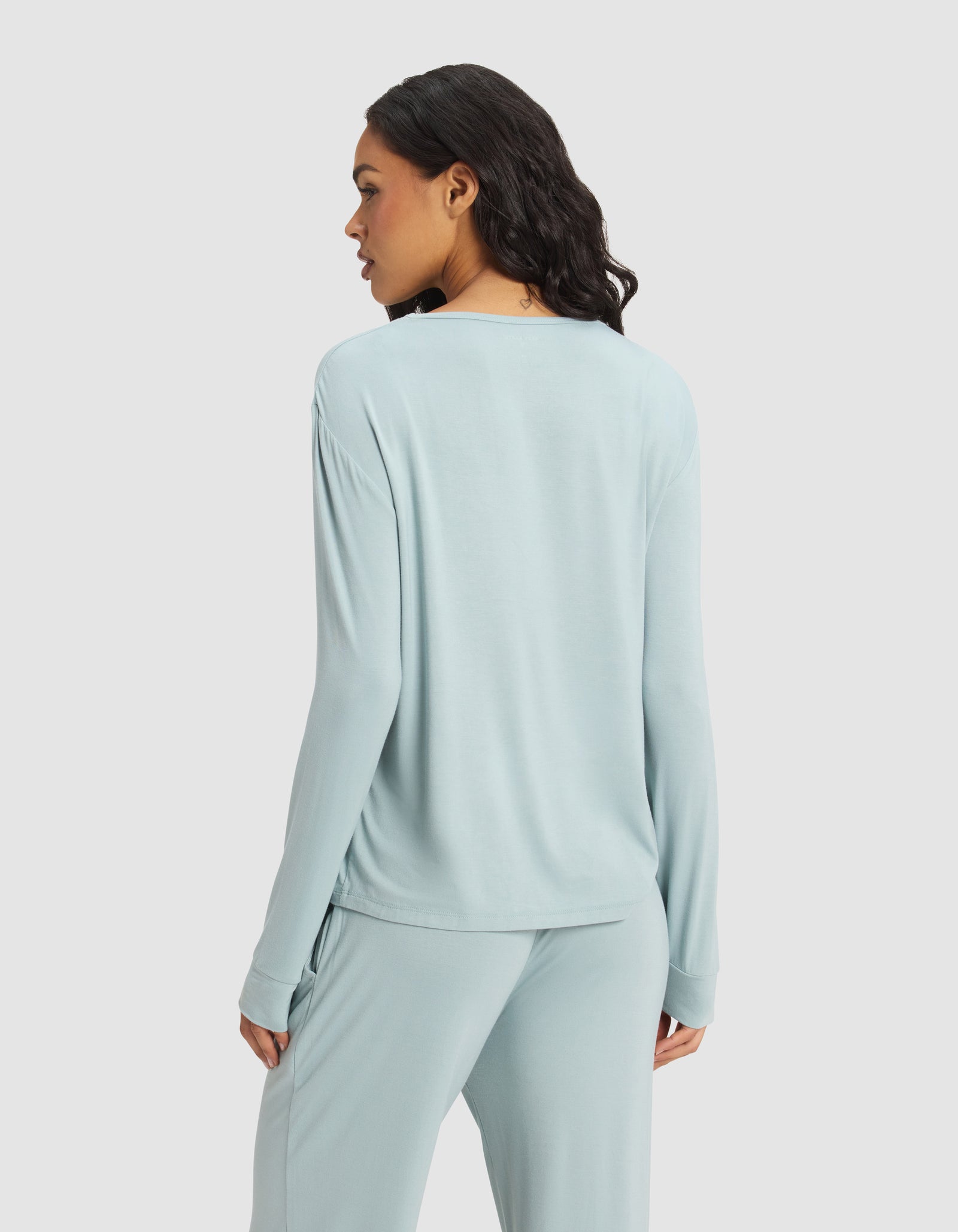 A woman with loose dark hair, seen from behind, wears a Cozy Earth Women's Bamboo Stretch-Knit Long Sleeve Lounge Tee in light blue and matching pants against a plain white background.