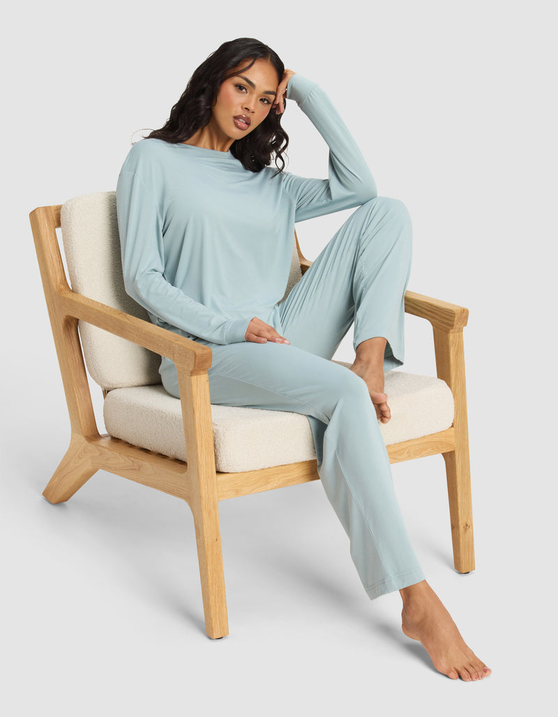 A woman relaxes on a cushioned wooden chair, wearing the Cozy Earth Women's Bamboo Stretch-Knit Long Sleeve Lounge Tee in light blue. One leg is bent and her arm rests on the chair's back. The background is plain and light-colored.