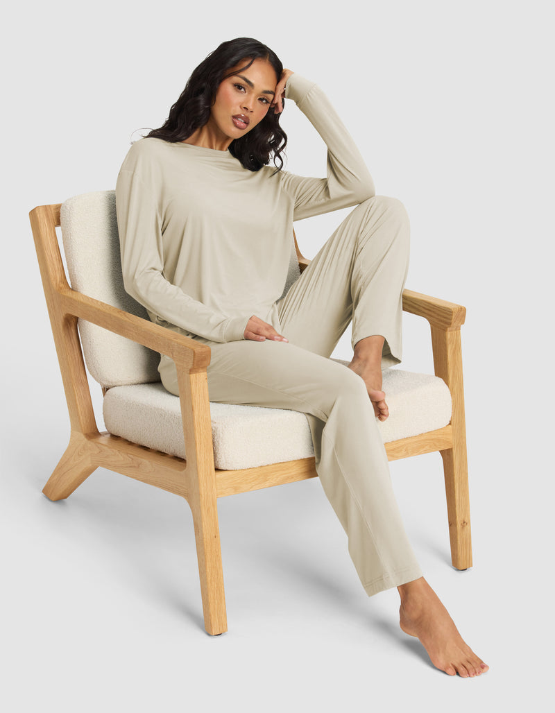 A woman relaxes barefoot in a wooden armchair with cream cushions, wearing the Cozy Earth Women's Loews Hotel Lounge Set—a light beige long-sleeve top and matching pants—with one arm resting on her head.