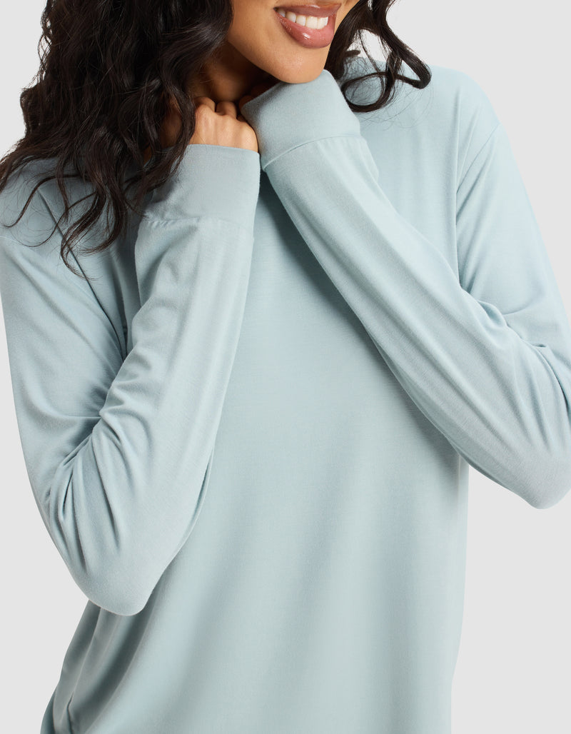 A woman with wavy dark hair smiles while holding the collar of her light blue Women's Bamboo Stretch-Knit Long Sleeve Lounge Tee by Cozy Earth, her face partially visible against a plain background.