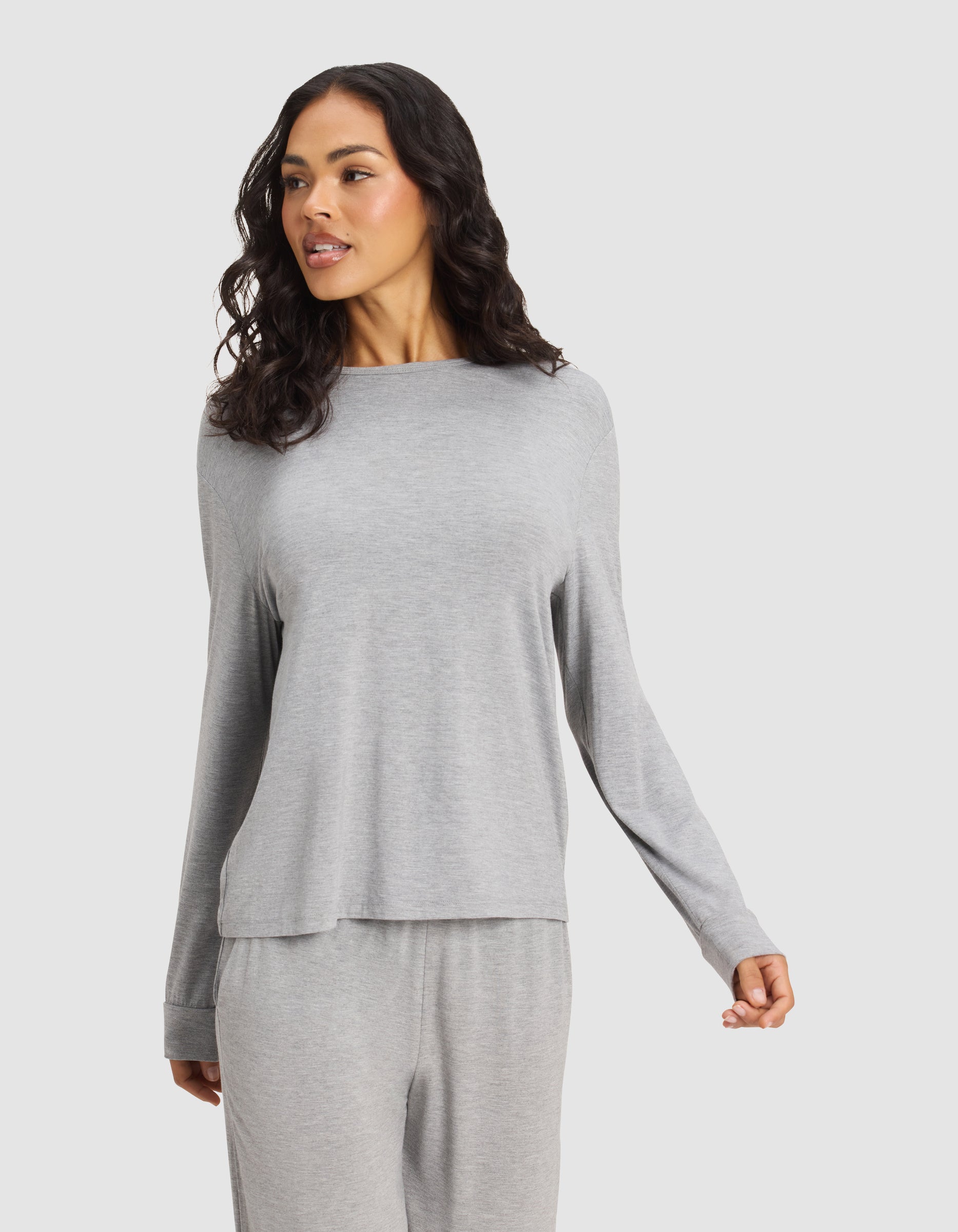 A woman with long, wavy dark hair models the Cozy Earth Women's Bamboo Stretch-Knit Long Sleeve Lounge Tee in light gray, paired with matching pants. She stands against a plain background, looking to the side with a relaxed expression.|Color:Grey