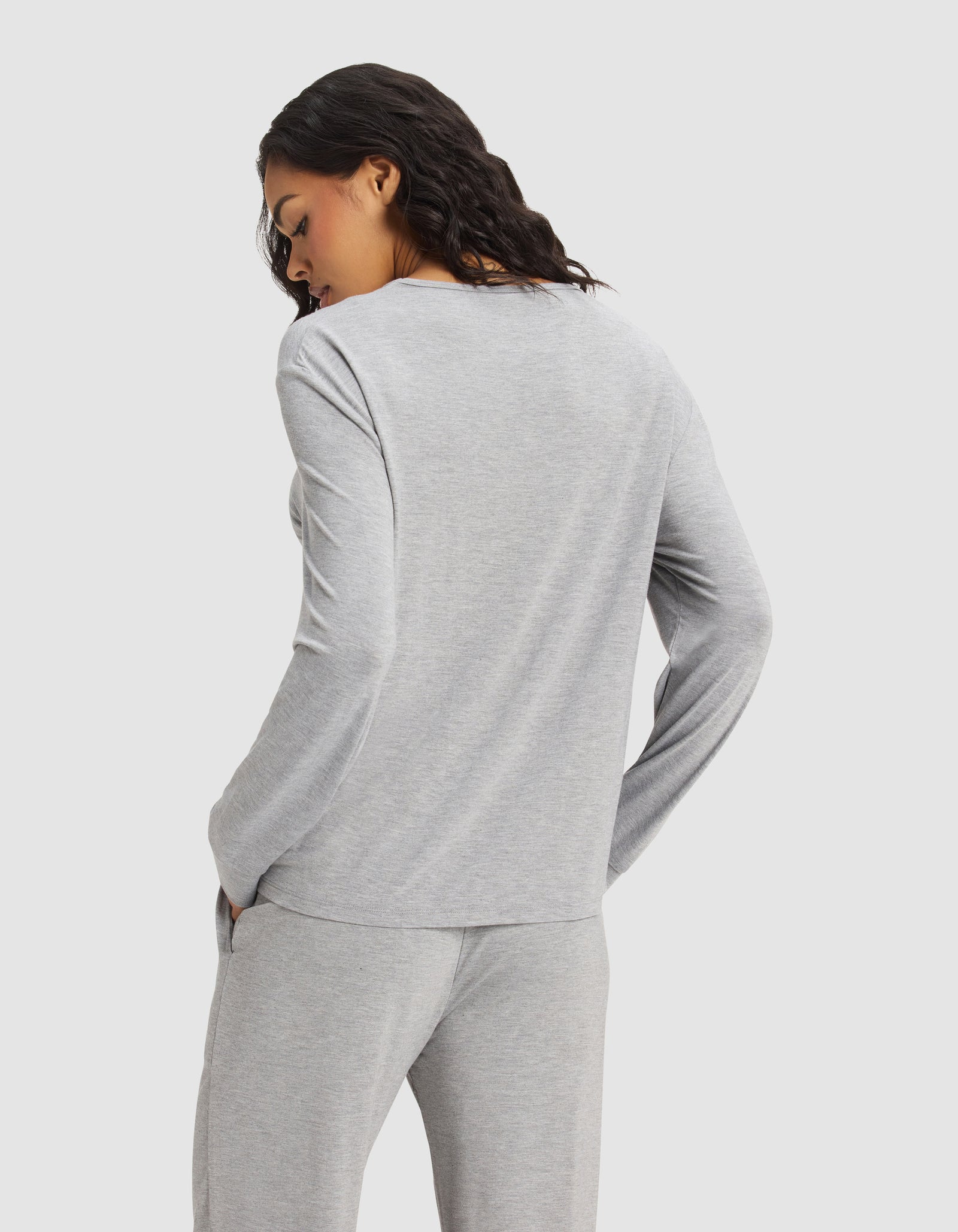 A woman with wavy dark hair is seen from behind, wearing the Cozy Earth Women's Bamboo Stretch-Knit Long Sleeve Lounge Tee in light gray, hands in her pockets, standing against a plain white background.