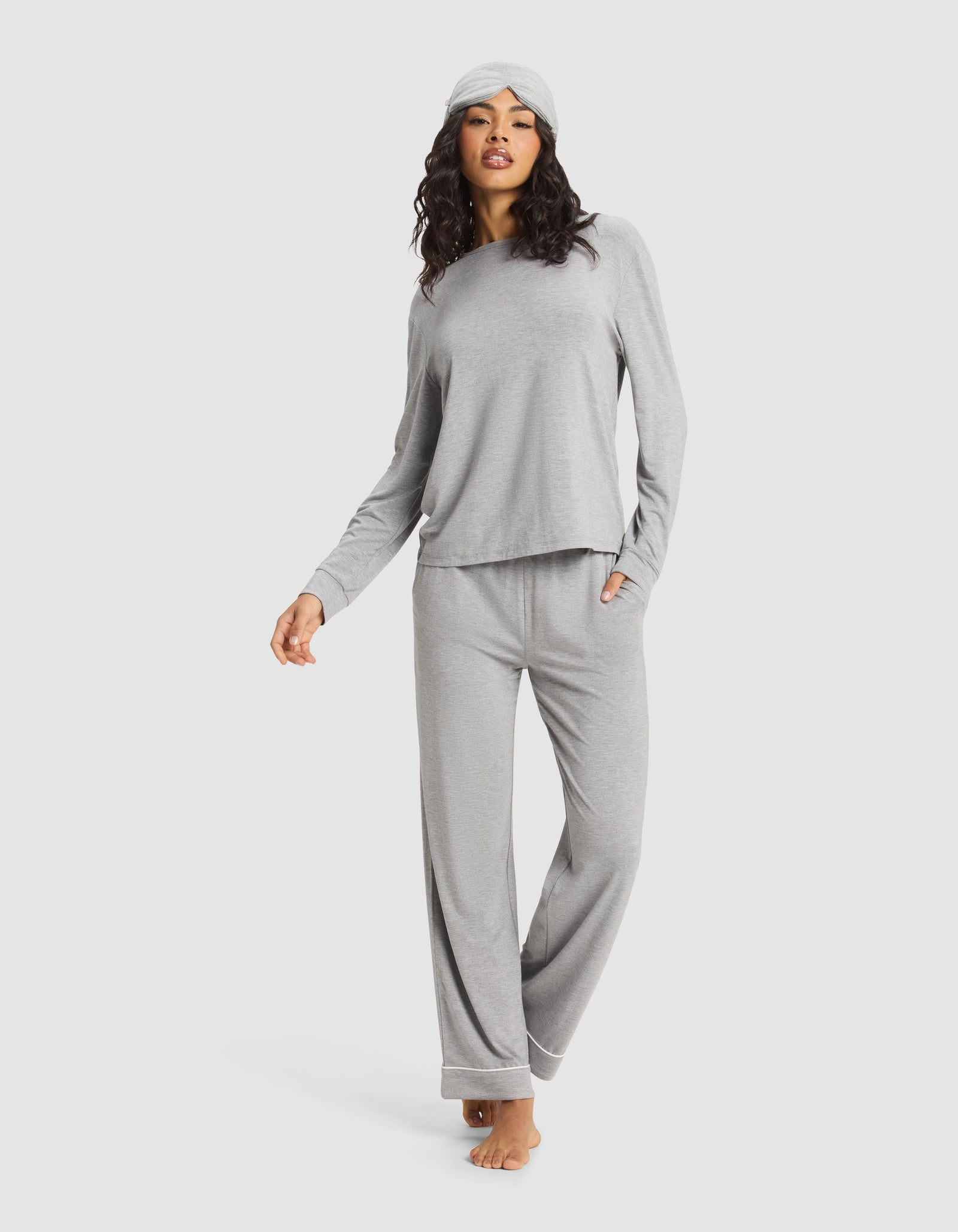 A woman stands barefoot against a white background, wearing Cozy Earth's Women's Bamboo Stretch-Knit Long Sleeve Lounge Tee in light gray with matching wide-leg pants and a sleep mask.