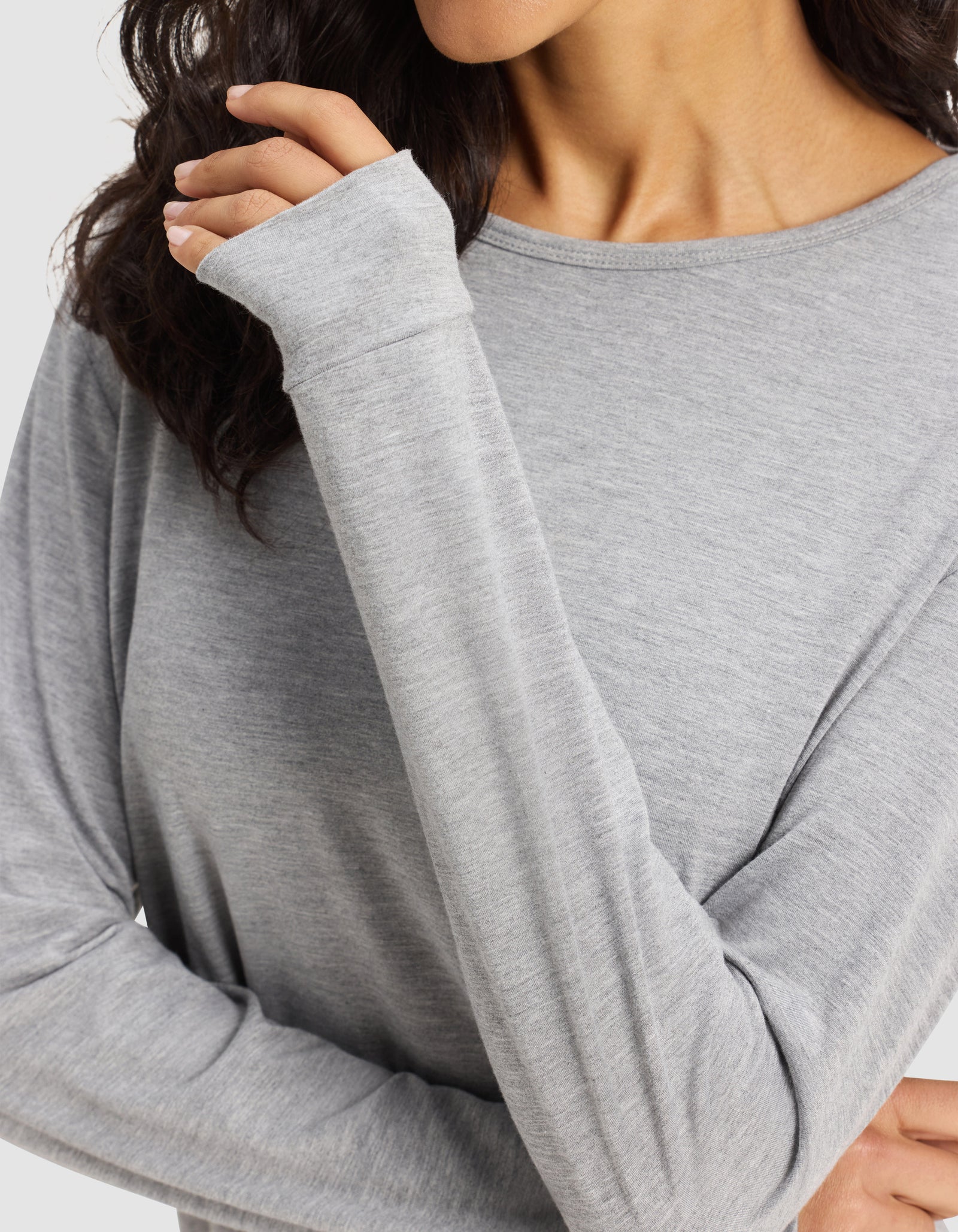 A woman wears the Cozy Earth Women's Bamboo Stretch-Knit Long Sleeve Lounge Tee in light gray, featuring thumb holes, with her left hand near her face and wavy dark hair partly visible.
