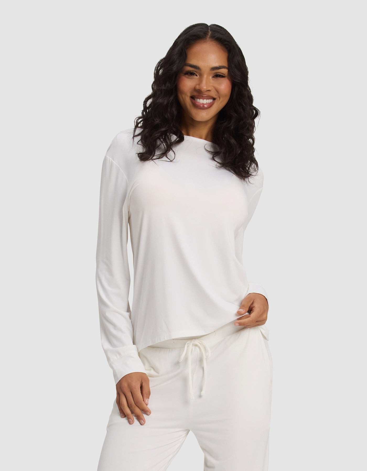 A woman with long, wavy dark hair smiles while wearing the Cozy Earth Women's Bamboo Stretch-Knit Long Sleeve Lounge Tee in white, paired with matching drawstring pants against a plain light gray background.