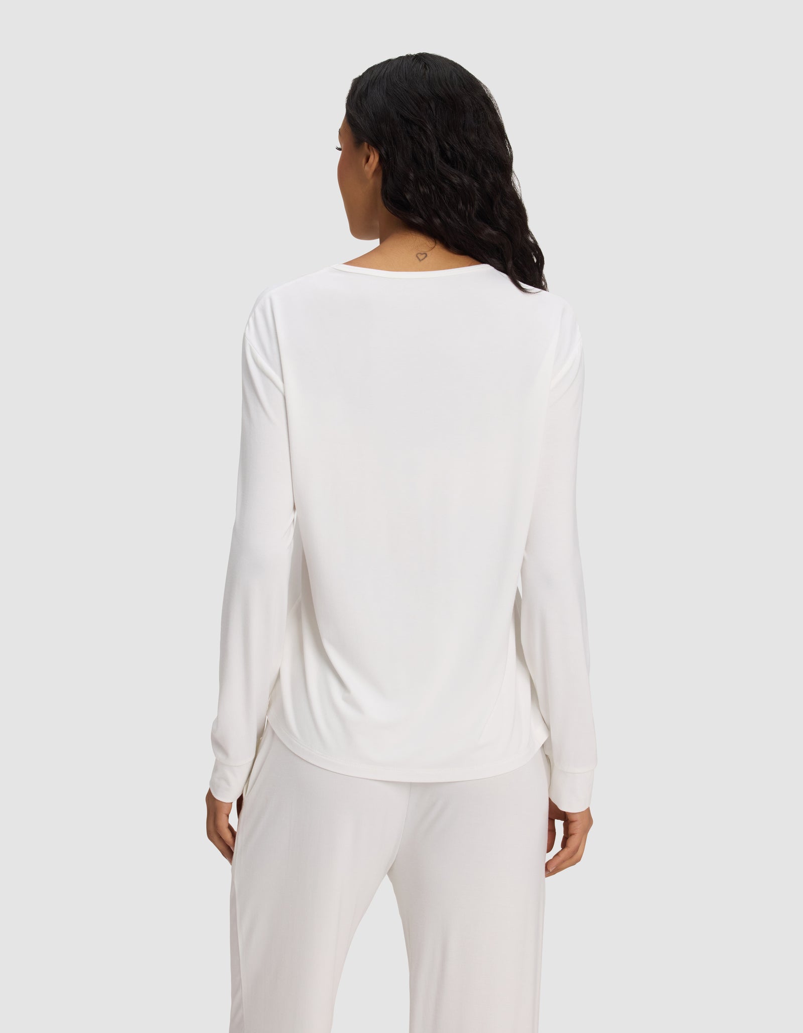 A woman with long dark hair is seen from the back, wearing the Cozy Earth Women's Bamboo Stretch-Knit Long Sleeve Lounge Tee in white, against a plain light gray background.