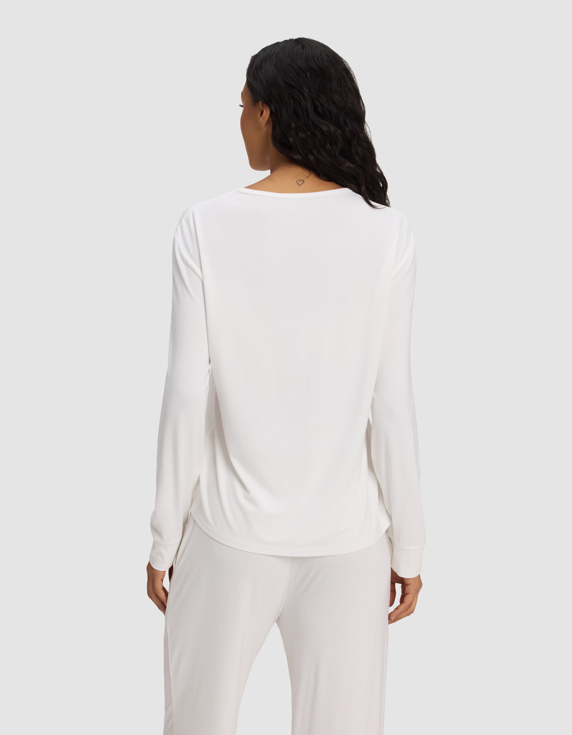 A woman with long dark hair is seen from the back, wearing the Cozy Earth Women's Bamboo Stretch-Knit Long Sleeve Lounge Tee in white, against a plain light gray background.